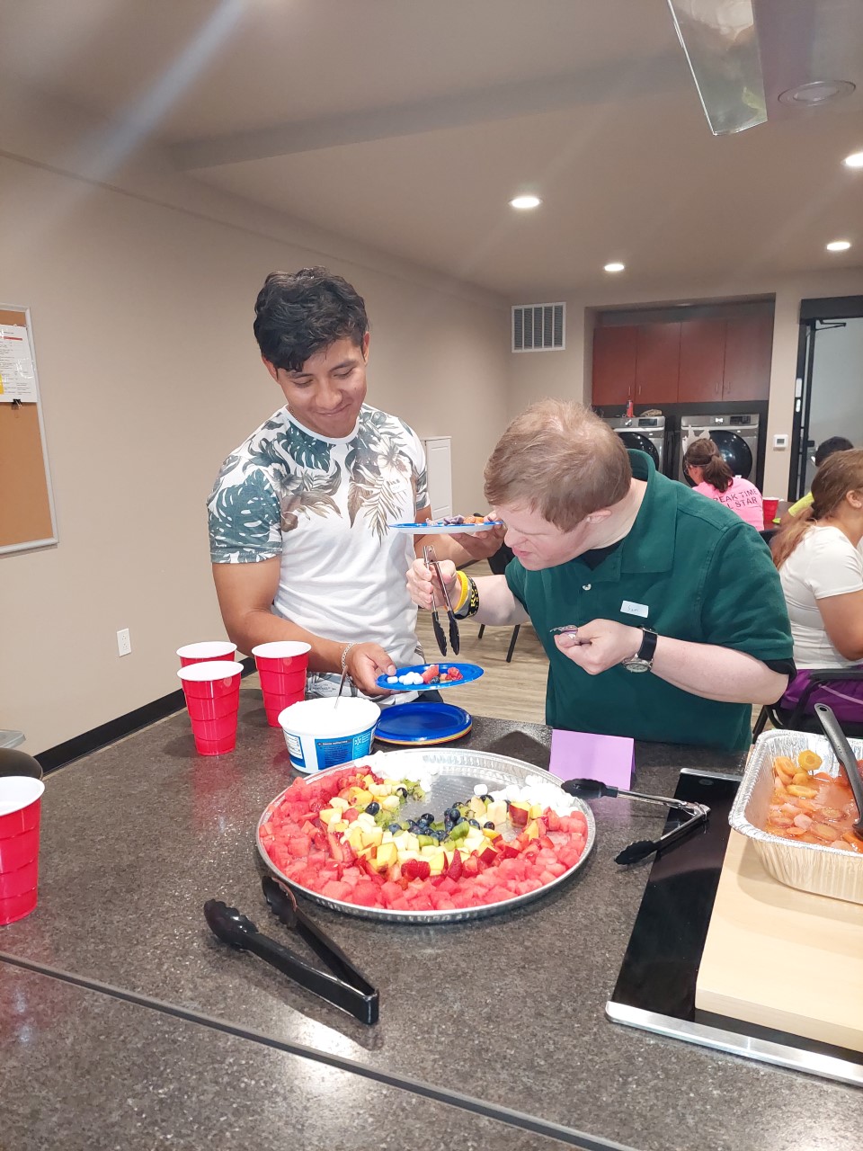 young adults getting food from a table including fruit at a gathering indoors