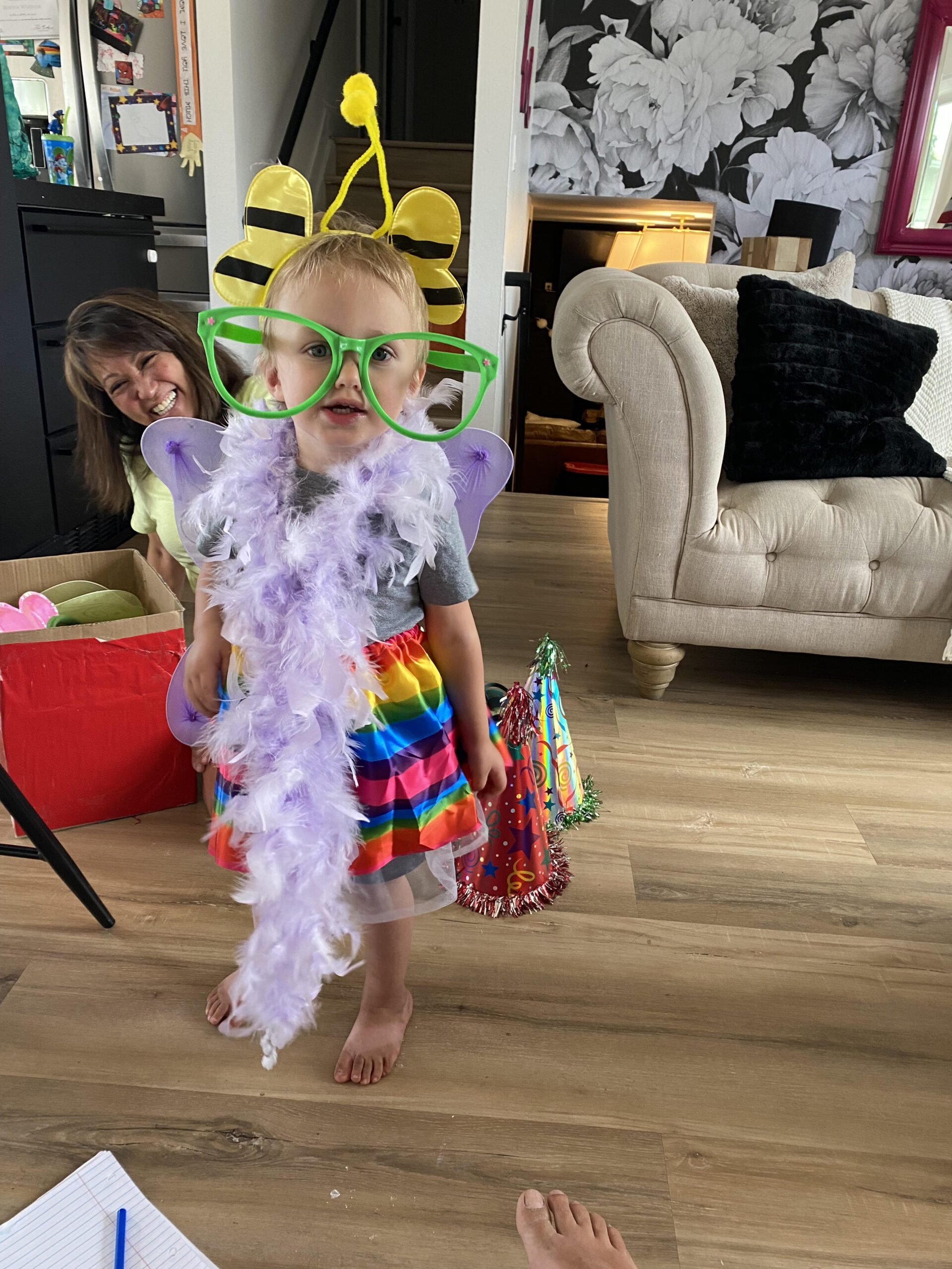 toddler looking at the viewer while wearing large green sunglasses, purple boa, bumblebee wings, and multi-colored skirt