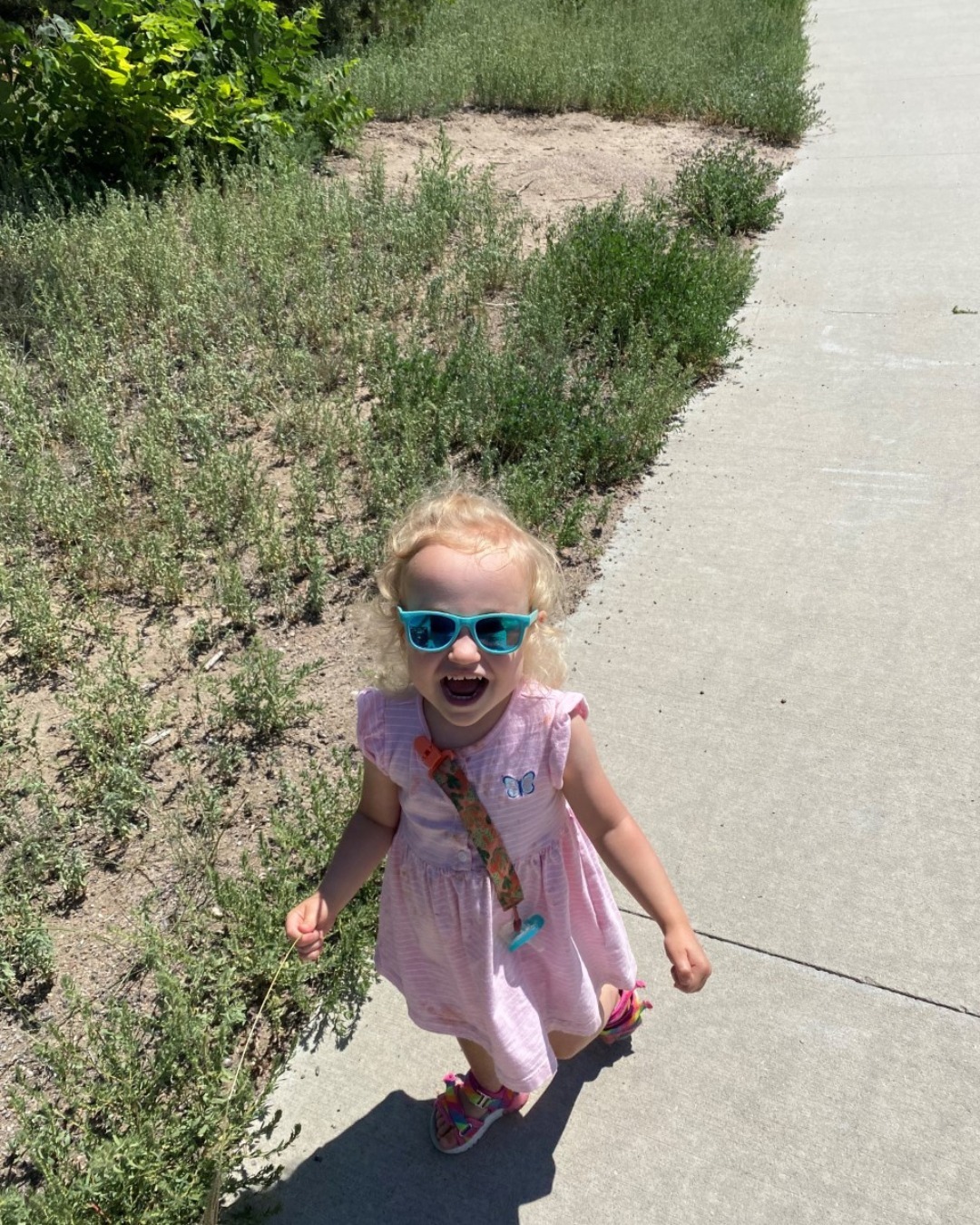 toddler wearing sunglasses smiling at viewer while walking down sidewalk on bright sunny day
