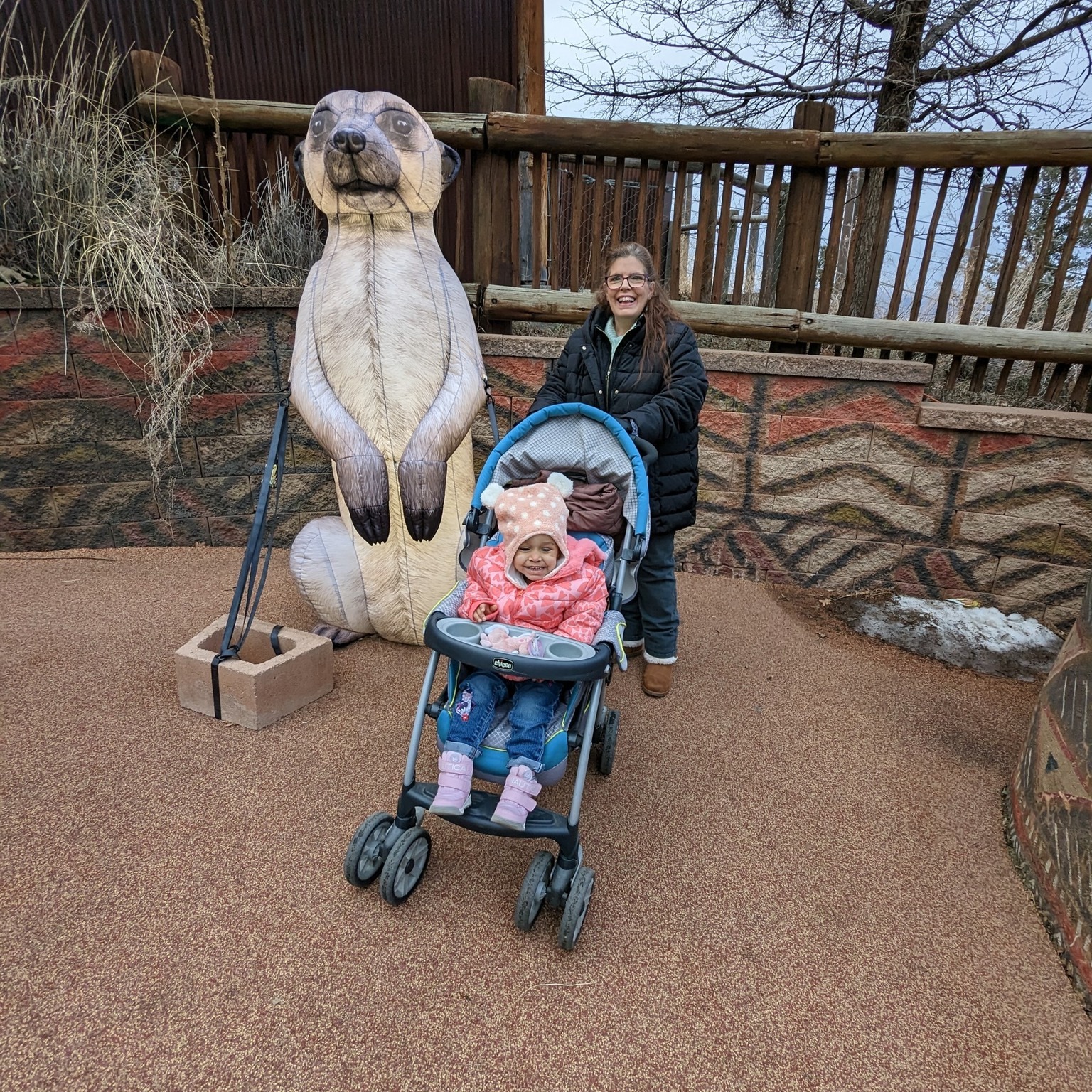 toddler seated in a stroller smiles at viewer while adult stands behind; they are both in front of a large plastic meerkat