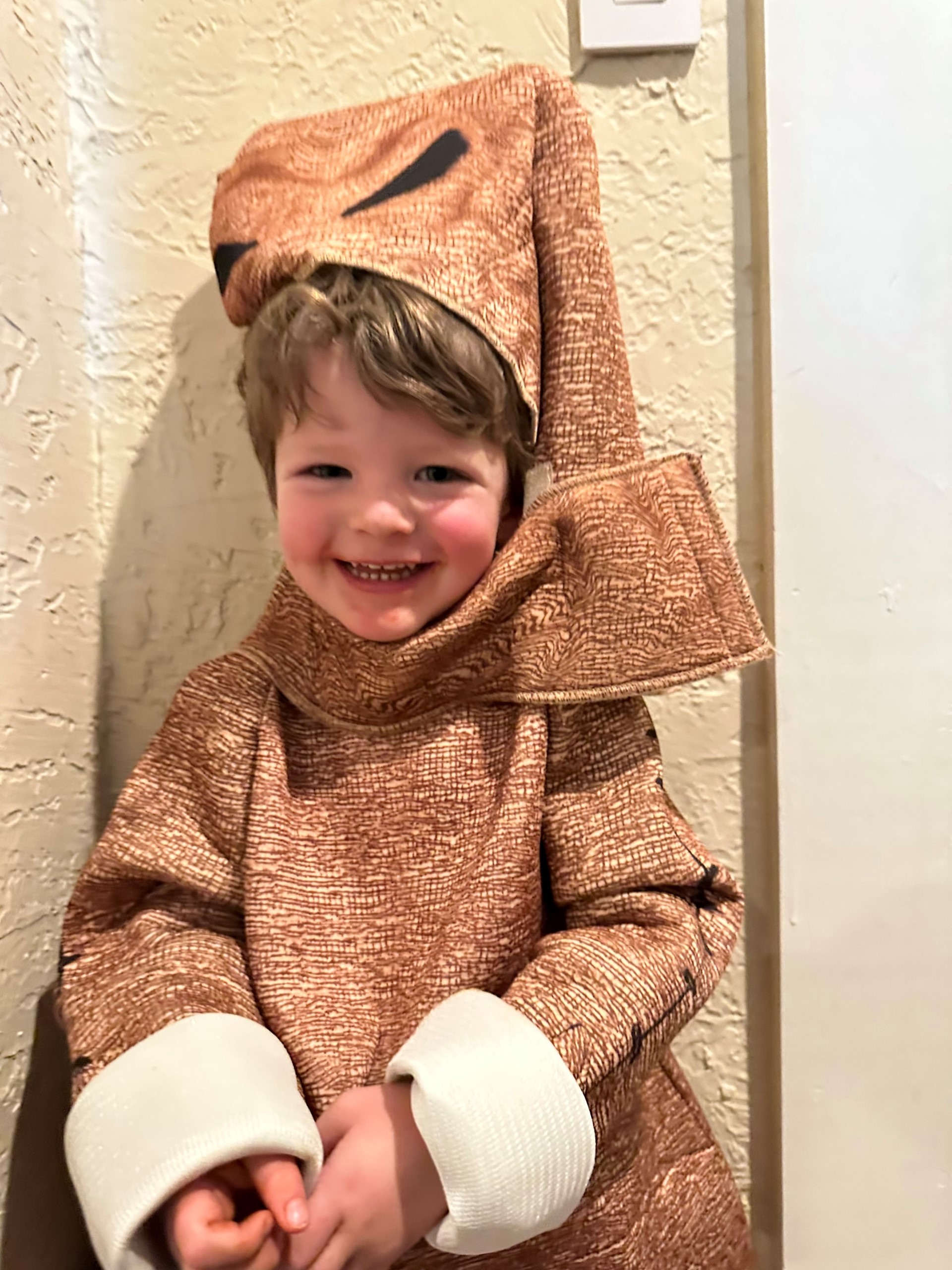 toddler smiling at the viewer wears a brown elvish costume with large white cuffs