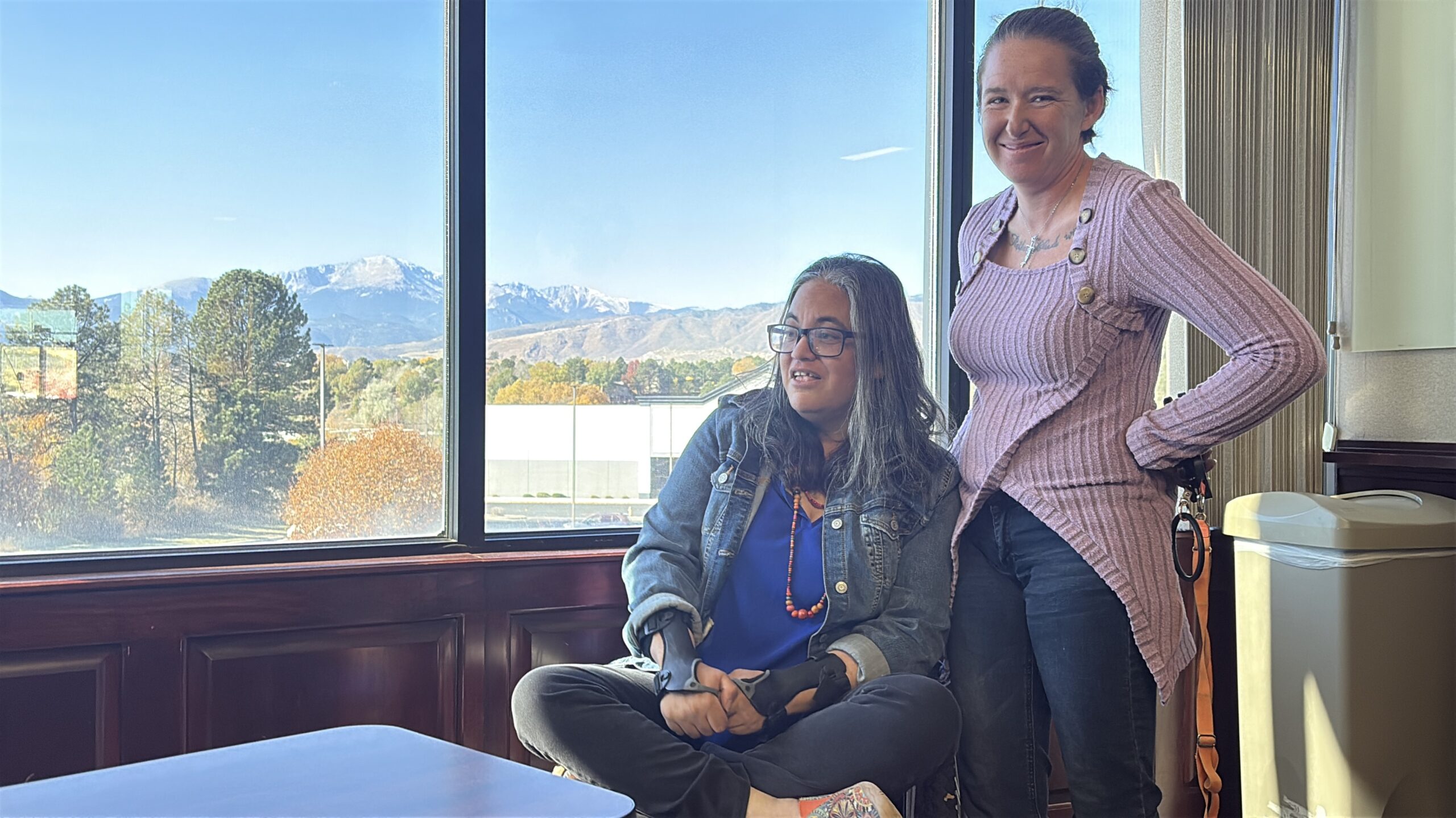 Sheera sits in her wheelchair with her legs crossed in front of a window with Pikes Peak behind her and Tashina standing next to her