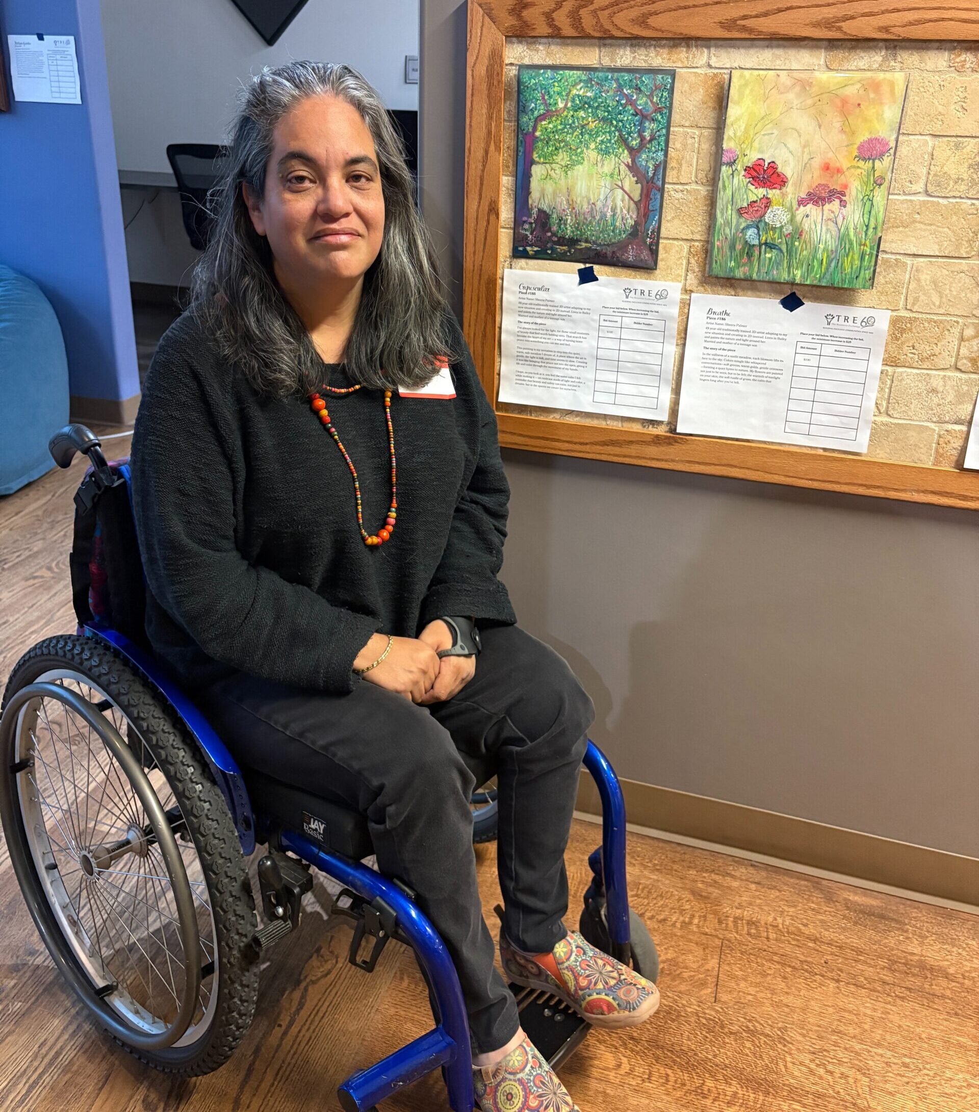 Sheera sits in her wheelchair in front of her two pieces of artwork on display 