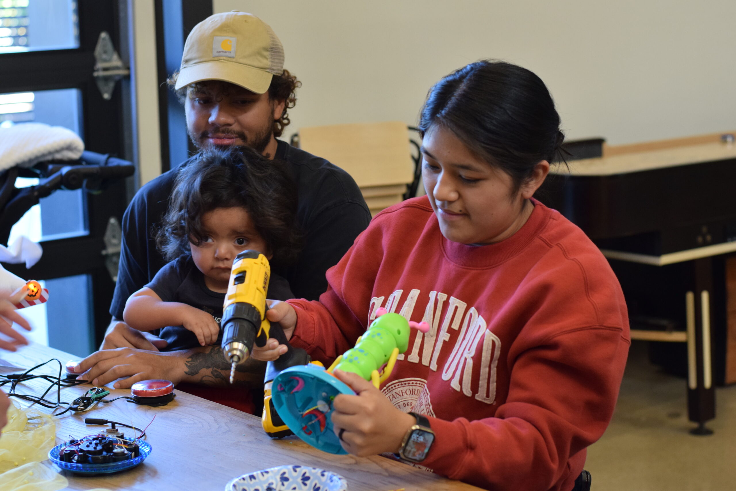 One parent drills into a toy in order to adapt it for accessibility with the other parent nearby holding their child with a disability
