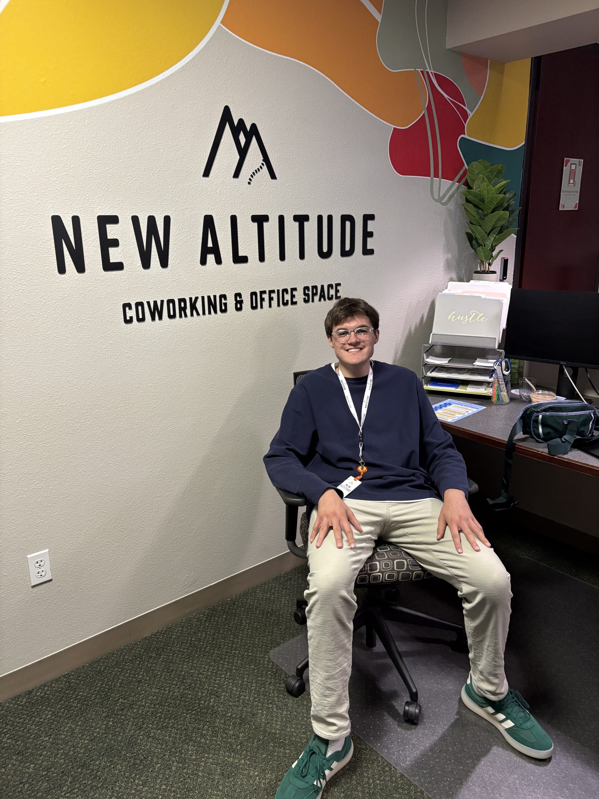 New Altitude Intern Caden sits behind the front desk in front of his computer with the New Altitude Coworking and Office Space logo behind him